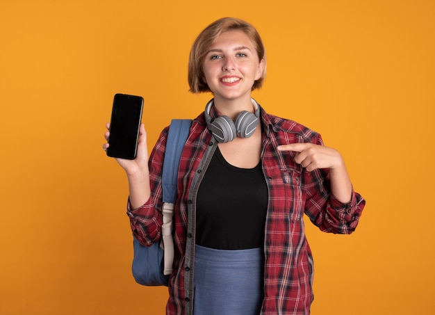 Jovem estudante eslava sorridente com fones de ouvido usando mochilas e aponta para o telefone