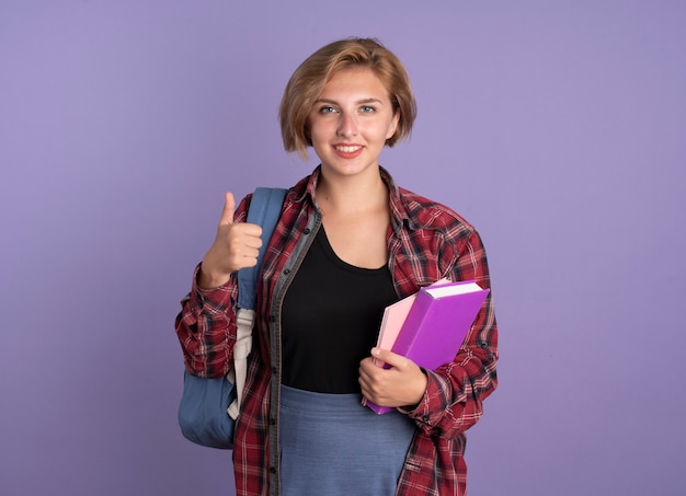 Jovem estudante eslava sorridente usando mochila com os polegares para cima segura o livro e o caderno