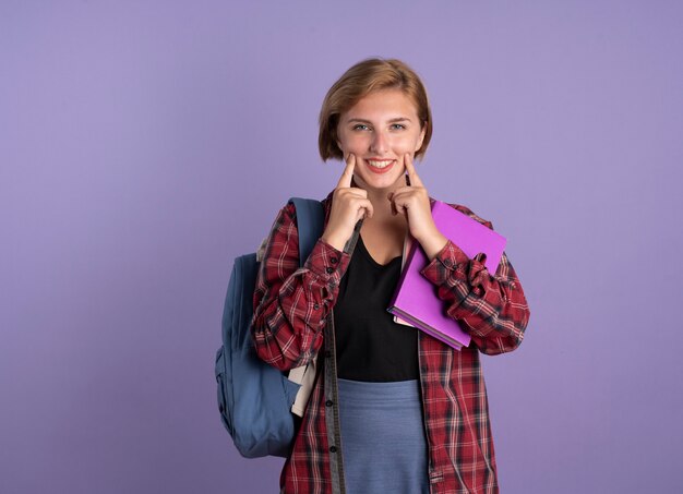 Jovem estudante eslava sorridente usando mochila continua sorrindo com os dedos segurando um livro e um caderno