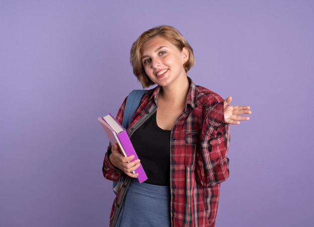 Jovem estudante eslava sorridente usando mochila mantém a mão aberta segurando livro e caderno