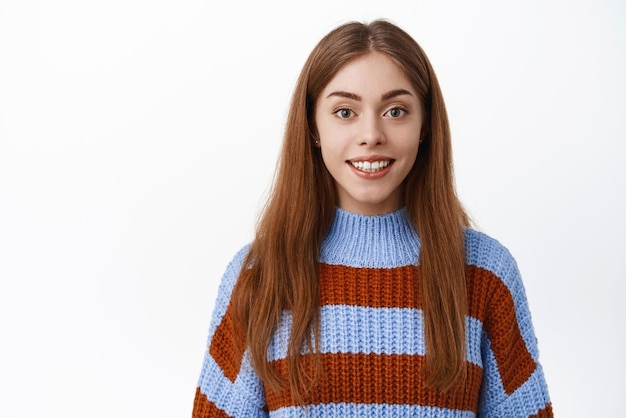 Jovem estudante feliz com cabelo comprido maquiagem natural de pé no suéter e sorrindo com fundo de estúdio de sorriso branco perfeito