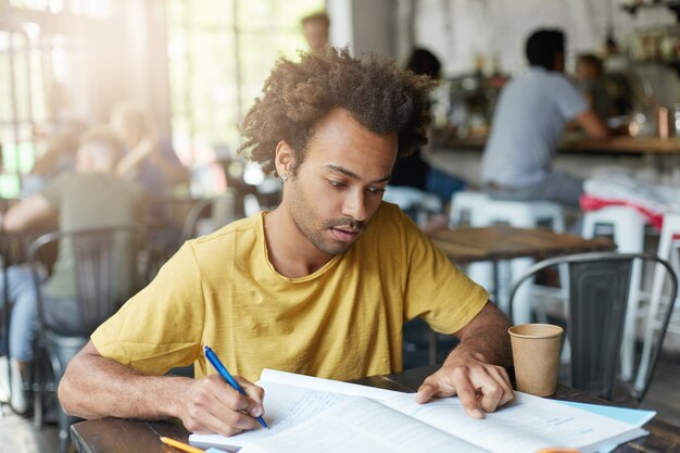 Jovem estudante negro, vestido casualmente, com barba e cabelos cacheados, tendo um olhar concentrado enquanto lê informações no livro e fazendo anotações no caderno, preparando-se para a aula na faculdade