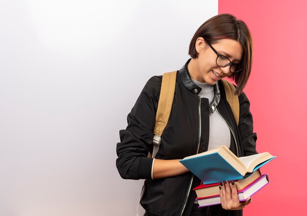 Jovem estudante sorridente usando óculos e bolsa traseira segurando e olhando para livros em frente a uma parede branca isolada na parede rosa