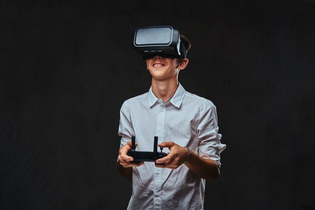 Jovem estudante vestido com uma camisa branca usando óculos de realidade virtual e detém um controle remoto quadcopter. Isolado em um fundo escuro.