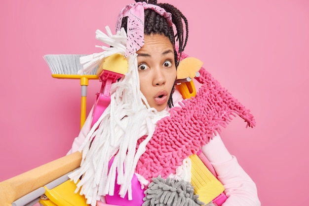 Foto grátis jovem estupefata chocada sobrecarregada com ferramentas de limpeza tem muito trabalho sobre a casa pronta para arrumar poses de apartamento contra fundo rosa conceito de limpeza e limpeza de primavera