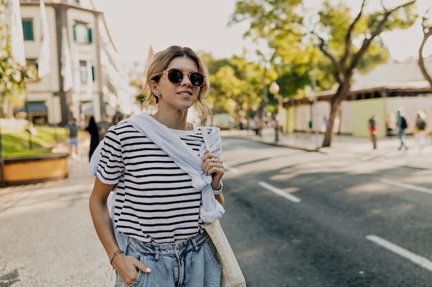 Jovem europeia com cabelo coletado vestindo camiseta listrada e óculos de sol está de pé na rua ensolarada no fundo de árvores exóticas e edifícios antigos