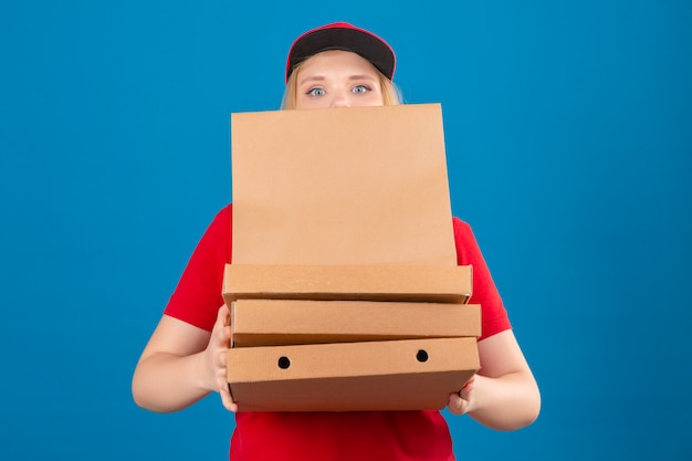 Jovem, exausta, entregadora, vestindo uma camisa pólo vermelha e boné em pé com uma pilha de caixas de pizza e um pacote de papel sobre um fundo azul isolado