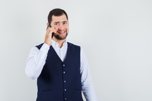 Jovem falando ao celular, de camiseta e colete, parecendo alegre