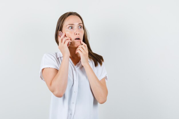 Jovem falando ao telefone de blusa branca e parecendo preocupada