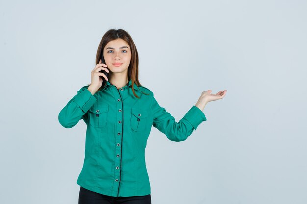 Jovem falando ao telefone, espalhando a palma da mão para o lado na blusa verde, calça preta e parecendo alegre, vista frontal.