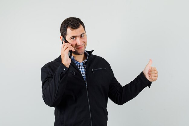 Jovem falando no celular, aparecendo o polegar na camisa, jaqueta e parecendo feliz, vista frontal.