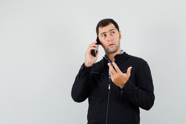 Jovem falando no celular, com camisa, jaqueta e parecendo hesitante. vista frontal.