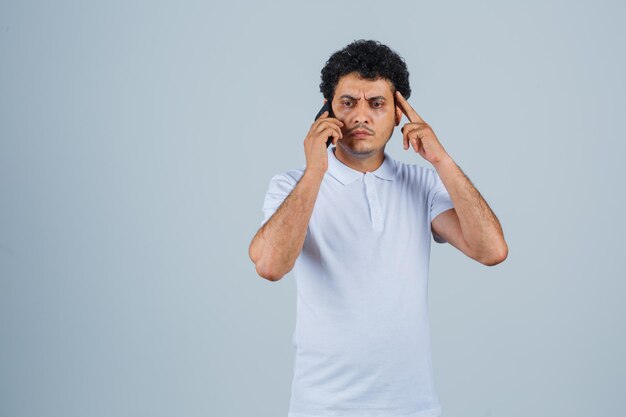 Jovem falando no celular em camiseta branca e parecendo esquecido. vista frontal.