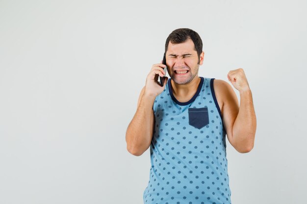 Jovem falando no celular, mostrando o gesto do vencedor em camiseta azul e olhando bem-aventurada, vista frontal.