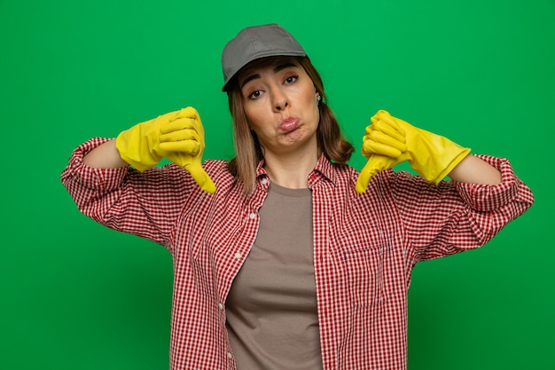 Jovem faxineira com camisa xadrez e boné usando luvas de borracha, olhando para a câmera descontente, mostrando os polegares para baixo em pé sobre um fundo verde