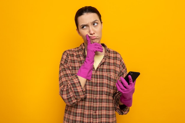 Jovem faxineira com camisa xadrez e luvas de borracha segurando um smartphone e olhando para o lado com uma expressão pensativa no rosto