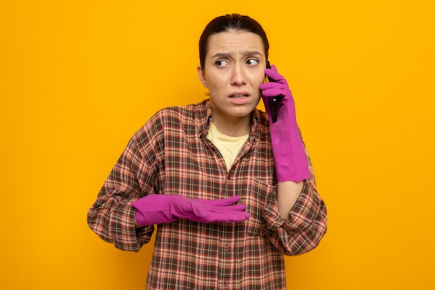 Jovem faxineira com roupas casuais e luvas de borracha, parecendo confusa enquanto fala no celular, em pé sobre a parede laranja