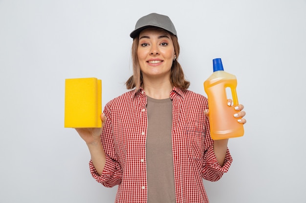 Foto grátis jovem faxineira em camisa xadrez e boné segurando uma garrafa de material de limpeza e uma esponja olhando para a câmera sorrindo alegremente feliz e positiva em pé sobre um fundo branco