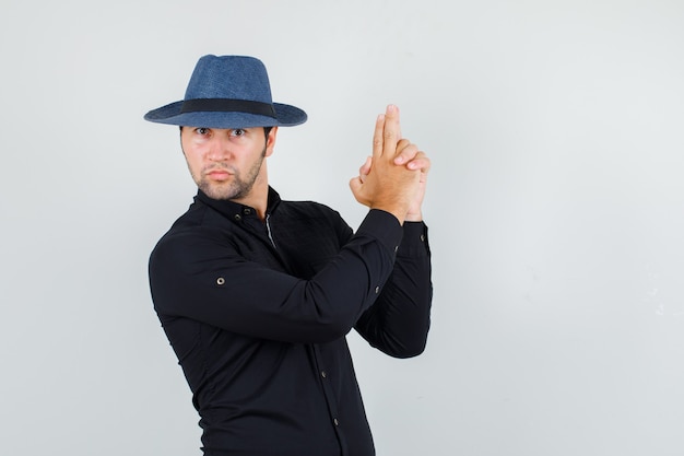 Foto grátis jovem fazendo gesto de pistola com as mãos na camisa preta, chapéu e parecendo rigoroso.