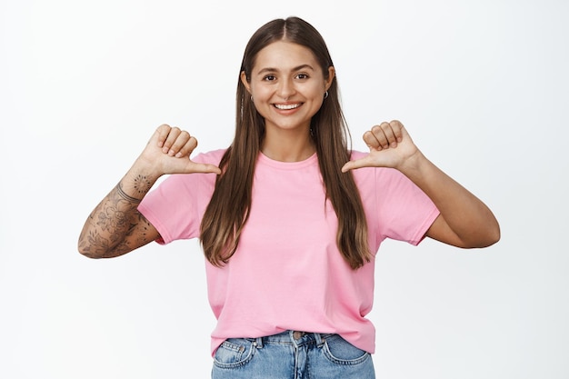 Foto grátis jovem feliz apontando o dedo para si mesma, mostrando o logotipo na camiseta rosa e sorrindo, de pé sobre fundo branco