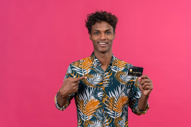 Jovem feliz bonito de pele escura com cabelo encaracolado, camisa estampada de folhas apontando para o cartão de crédito com o dedo indicador