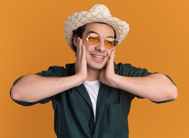 Jovem feliz com camisa verde e chapéu de verão, usando óculos olhando para o lado com um sorriso no rosto em pé sobre uma parede laranja