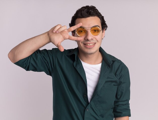 Jovem feliz com camisa verde, usando óculos, olhando para a frente, sorrindo alegremente, mostrando o sinal V perto dos olhos, em pé sobre uma parede branca