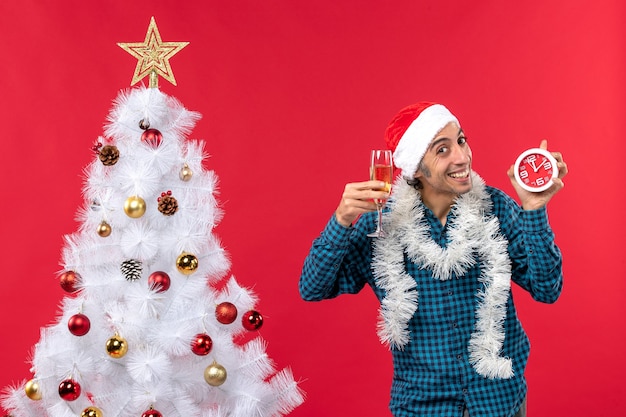 Jovem feliz com chapéu de papai noel e segurando uma taça de vinho e um relógio perto da árvore de natal no vermelho