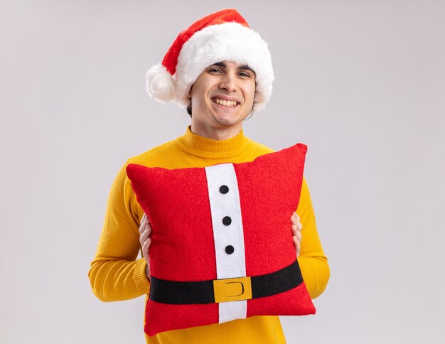 Jovem feliz com gola alta amarela e chapéu de Papai Noel segurando a almofada de Natal, olhando para a câmera com um sorriso no rosto em pé sobre um fundo branco
