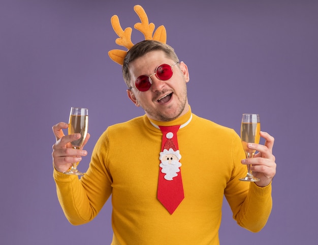 Jovem feliz com gola alta amarela e óculos vermelhos, gravata vermelha engraçada e aro com chifres de veado, segurando duas taças de champanhe com um grande sorriso no rosto em pé