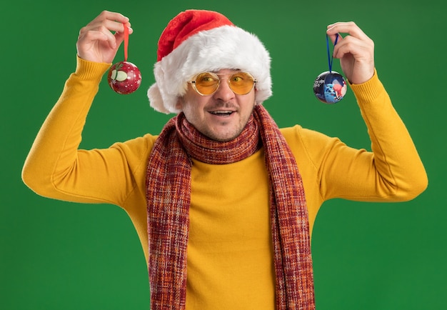 Jovem feliz com gola amarela e chapéu de papai noel usando óculos segurando brinquedos para a árvore de natal, olhando para a câmera sorrindo alegremente em pé sobre um fundo verde