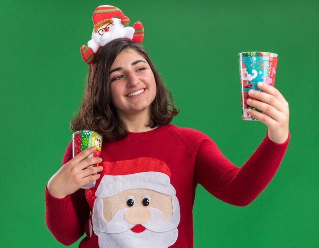 Foto grátis jovem feliz com suéter de natal com chapéu de papai noel segurando um copo de papel colorido olhando para ele com um sorriso no rosto em pé sobre a parede verde