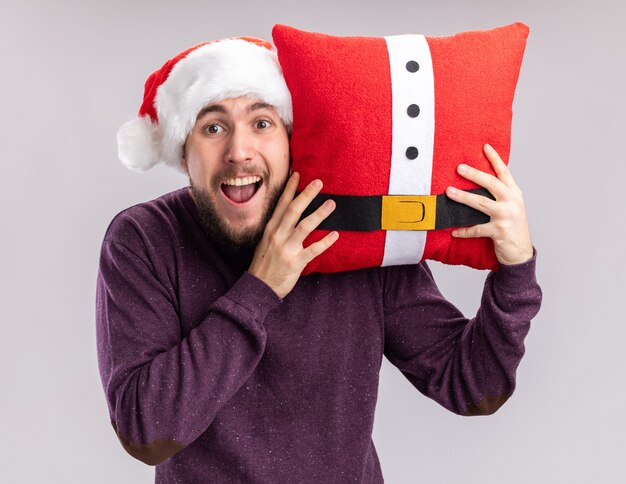 Jovem feliz com suéter roxo e chapéu de Papai Noel segurando uma almofada engraçada, olhando para a câmera espantado e surpreso em pé sobre um fundo branco