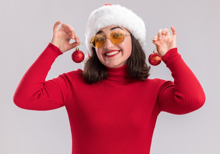Foto grátis jovem feliz com suéter vermelho e chapéu de papai noel usando óculos segurando bolas de natal, olhando para a câmera, sorrindo amplamente em pé sobre um fundo branco