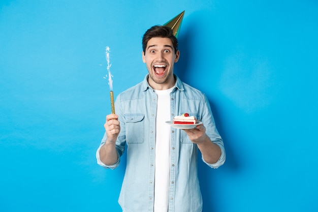 Jovem feliz comemorando aniversário com chapéu de festa, segurando um bolo de aniversário e sorrindo, em pé sobre um fundo azul