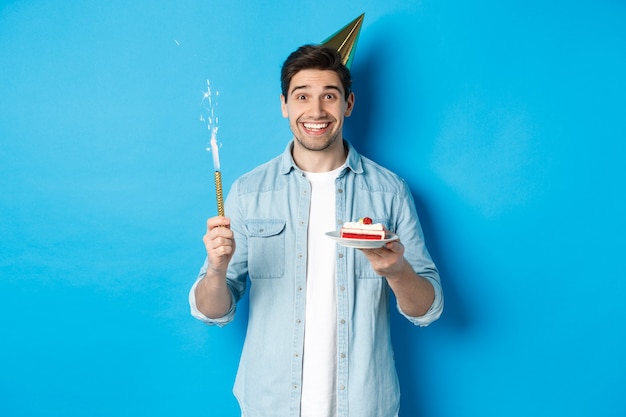 Jovem feliz comemorando aniversário com chapéu de festa, segurando um bolo de aniversário e sorrindo, em pé sobre um fundo azul