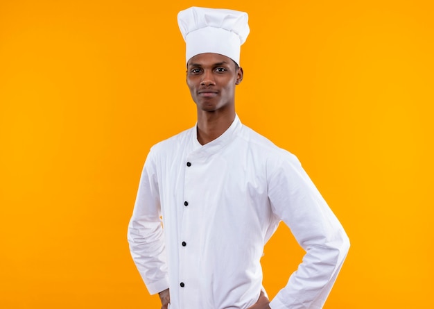 Jovem feliz cozinheira afro-americana com uniforme de chef coloca as mãos na cintura isolada na parede laranja