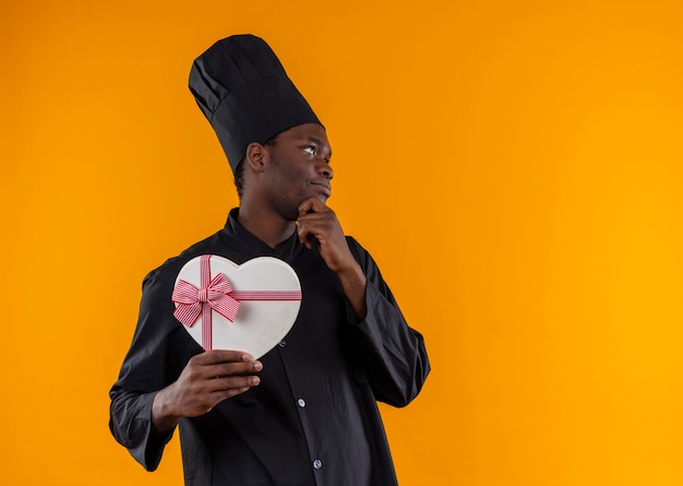 Jovem feliz cozinheira afro-americana com uniforme de chef segura uma caixa em formato de coração e coloca a mão no queixo em uma laranja com espaço de cópia