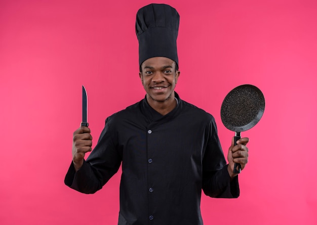 Jovem feliz cozinheira afro-americana com uniforme de chef segurando uma faca e uma frigideira isolada na parede rosa