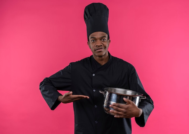 Jovem feliz cozinheira afro-americana com uniforme de chef segurando uma panela e segurando a mão reta isolada na parede rosa