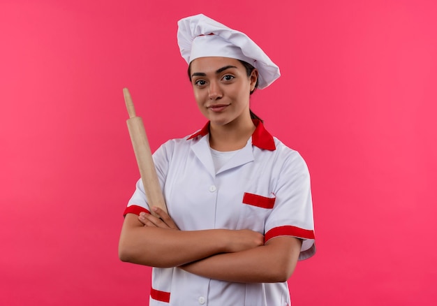 Jovem feliz cozinheira caucasiana com uniforme de chef cruza os braços e segura o rolo de massa isolado na parede rosa com espaço de cópia