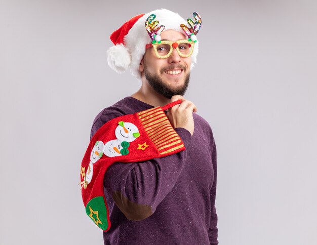 Jovem feliz e alegre com suéter roxo e chapéu de Papai Noel usando óculos engraçados, segurando uma meia de Natal, olhando para a câmera sorrindo amplamente em pé sobre um fundo branco