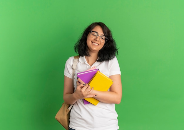 Jovem feliz e bonita caucasiana, usando óculos e bolsa traseira, segurando livros em verde com espaço de cópia