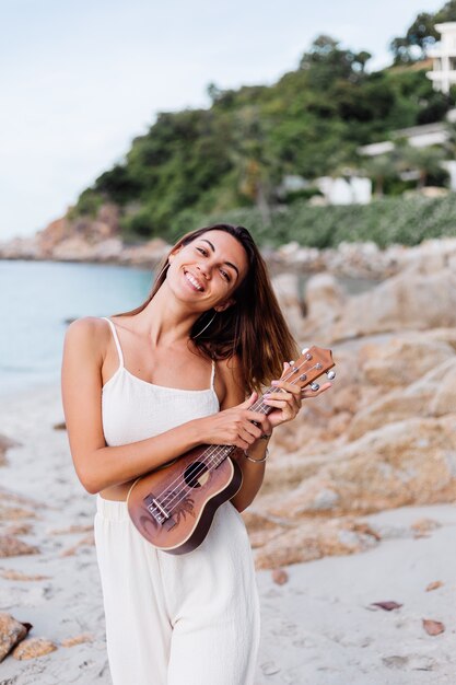 jovem feliz e calma mulher caucasiana com ukulele na praia tropical rochosa ao pôr do sol