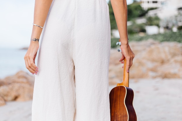 jovem feliz e calma mulher caucasiana com ukulele na praia tropical rochosa ao pôr do sol