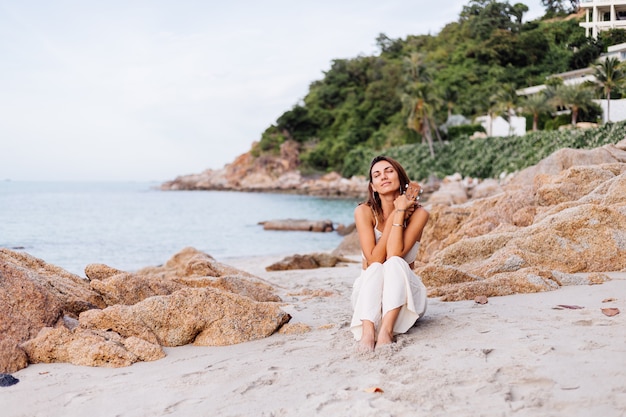 jovem feliz e calma mulher caucasiana com ukulele na praia tropical rochosa ao pôr do sol