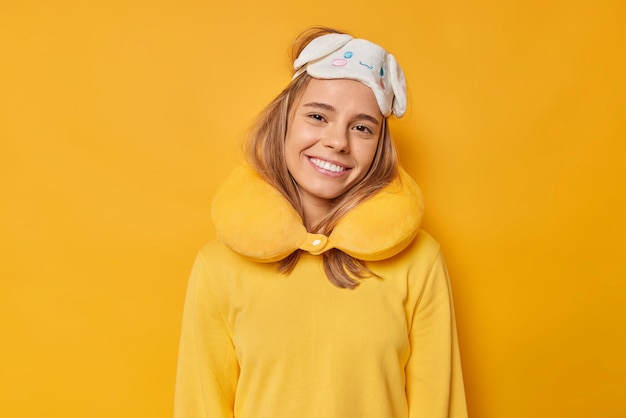 Jovem feliz e encantada mulher europeia com sorrisos dentuços, usa máscara de dormir no travesseiro de pescoço e está de bom humor, vestida com um macacão casual isolado sobre um fundo amarelo vívido se prepara para descansar