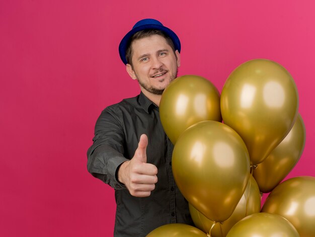 Jovem feliz e festeiro com um boné azul parado ao lado de balões, mostrando o polegar isolado em uma rosa