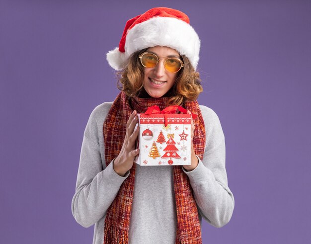 jovem feliz e positivo usando chapéu de Papai Noel de Natal e óculos amarelos com um lenço quente em volta do pescoço segurando um presente de Natal, olhando para a câmera com um sorriso no rosto em pé sobre um fundo roxo
