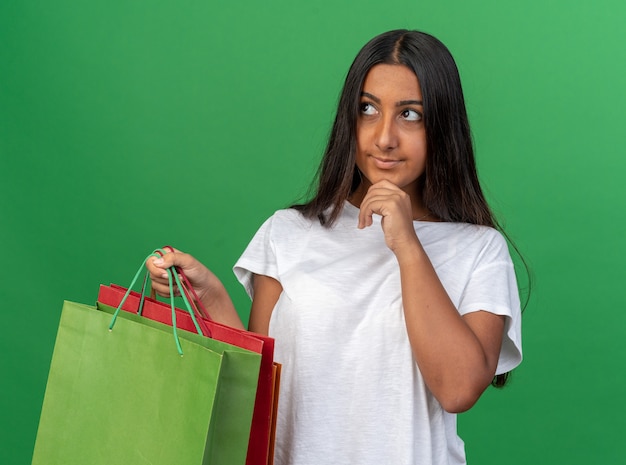 Jovem feliz em uma camiseta branca segurando sacolas de papel olhando para o lado com a mão no queixo e uma expressão pensativa pensando